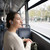 Person in cream jumper wearing blue wireless earbuds on a bus, window view in background