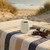 Portable white wireless speaker on striped towel at beach, with sunglasses and book