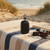Black portable Bluetooth speaker on striped towel at beach, next to sunglasses and a book