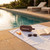 Portable wireless speaker, sunglasses, book and sunscreen on white towel by an outdoor pool