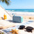 Blue portable Bluetooth speaker on beach towel beside sunglasses, book and sunscreen.
