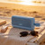 Blue portable Bluetooth speaker on sunlounger with sunglasses, sunscreen and beach in background