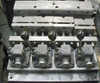 Four 2-FLs with fixturing. The knobs on the end of the center bars have been replaced by discs. A tool is used to pry back the center bar and v-blocks to load the part. The springs hold the part in place before the locking lever is closed. The dowels on either side of the v-block keep the v-block aligned properly.