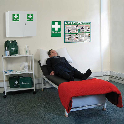 This image depicts a first aid room setup in a school environment. The room features a first aid bed with a red cushion, a first aid cabinet, and a first aid sign on the wall. A person is lying on the bed, and the room is equipped with the necessary supplies to assist in minor medical situations. A wall-mounted first aid poster is visible.