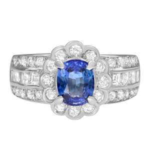 An image of a Rachel Koen women's ring with a central oval-cut blue sapphire surrounded by a halo of round-cut diamonds in platinum. The band is adorned with two rows of smaller round cut diamonds and princess cut diamonds set at a frontal angle, providing a clear and detailed view of the ring's design. The image is taken from a close distance, showcasing the sparkling facets of the blue sapphires and the intricate setting of the ring.