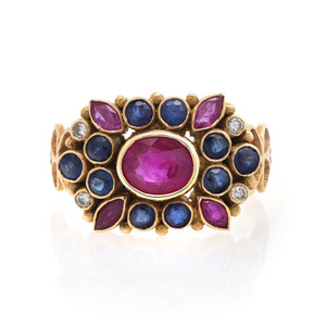 An image of a Rachel Koen women's cocktail ring with diamonds and Ruby and Blue Sapphire, displayed front-facing at a close distance. The ring features a prominent central oval Ruby, flanked by smaller and blue Sapphire in teardrop and round shapes, all set in a textured yellow gold band with interspersed diamond accents. The ring is photographed against a white background, with a slight reflection visible beneath it.