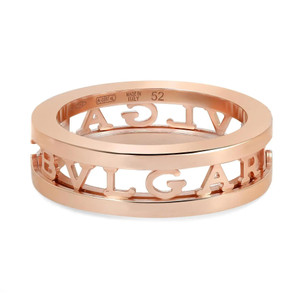 An image of a Bvlgari B.Zero1 Openwork Logo Spiral unisex adult one band ring without stones. The ring is captured in close-up, occupying the center of the frame, and displayed at a slight angle to show its three-dimensional design. The ring has a rose gold finish and features the iconic Bvlgari logo cut out along its circumference. The inner band is visible, and engraving indicating the brand, size, and 'Made in Italy' can be seen. The background is a neutral, light color that contrasts with the warm tones of the ring. The condition of the ring is new. 