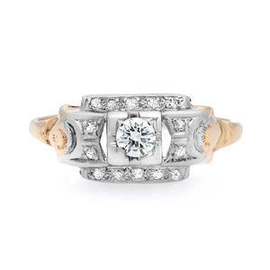 An image of a Rachel Koen brand women's antique engagement ring with diamonds, showcased in a front-facing view with a slight angle that allows for the visibility of the top and side details. The ring features a large central round cut diamond, surrounded by smaller diamonds set in a rectangular formation. Additional diamonds are embedded along the band, which has intricate designs and a two-tone gold scheme. The ring is positioned against a white background, and the shot is taken from a close, yet fully encompassing distance to capture the sparkle and design of the ring.The ring is pre-owned in a good condition. 