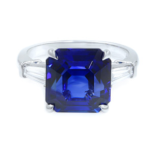 An image of a Rachel Koen women's engagement ring with diamonds, featuring a prominent asscher-cut sapphire center stone flanked by two clear, baguette side stones. The ring is displayed at a front-facing angle with the platinum band slightly visible, positioned centrally against a white background, offering a clear view of the gem settings and the intricate cuts of the stones. The distance is such that the ring fills the frame, allowing for detailed observation.