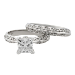 An image of a Rachel Koen women's bridal ring set with diamonds, a combination of an engagement ring and a wedding ring band. Engagement ring prominently featuring a large, princess-cut center diamond in a prong setting. The ring is captured in a close-up side view showing the band's intricate design with multiple smaller diamonds encrusted along its looped, overlapping structure. Both band exhibit a polished white gold finish, and the image is taken from a slight angle to highlight the sparkle of the diamonds and the details of the setting. The background is neutral, ensuring the focus remains on the ring's craftsmanship and brilliance.