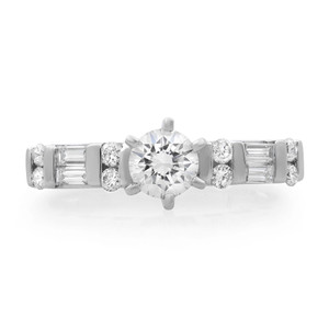 An image of a Rachel Koen brand engagement ring for women, featuring a prominent central round-cut diamond in a prong setting, flanked by a series of smaller round and baguette-cut diamonds set into the band. The ring is photographed in a direct frontal view against a white background, with the band parallel to the viewing angle, allowing for a clear view of the diamonds and the design details. The image is taken from a close distance, providing a detailed view of the ring's craftsmanship and sparkle.The ring is pre-owned in great condition. 