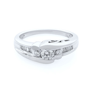 An image of a Rachel Koen brand women's engagement ring with diamonds, displayed at a slight angle to show the profile and top view. The ring features a central round brilliant cut diamond flanked by smaller round diamonds, and a row of channel-set baguette diamonds extending down the shoulders. The photo is taken from a close distance against a white background, with the focus on the diamonds and the polished white gold band.