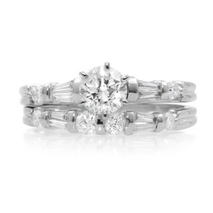 An image of a Rachel Koen brand women's engagement and bridal ring set with diamonds, displayed frontally at a close distance. The ring features a prominent round-cut diamond centered at the top, flanked by baguette-cut diamonds on its shoulders, with smaller round-cut diamonds set along the band. The band is shown horizontally in the image, with the main diamond facing the viewer and the ring's intricate details clearly visible due to the angle and lighting. The ring is pre-owned in a great condition. 