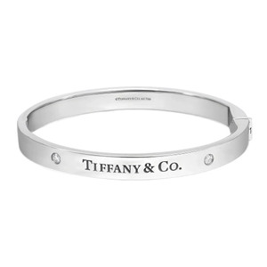 An image of a Tiffany & Co. unisex adult bracelet with diamonds. The bracelet is shown in a close frontal view, with a slight angle to display its curvature and thickness. It features a polished white gold band with the iconic Tiffany & Co. brand name engraved in the center. Two small, round-cut diamonds are symmetrically set on the band, adding a touch of sparkle. The image background is white, emphasizing the bracelet's design and shine.