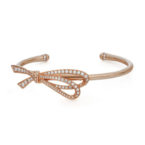 An image of a women's Tiffany & Co. bracelet with diamonds, presented on a white background. The bracelet is in a cuff style in rose gold, featuring a decorative bow design at the top. The bow is adorned with numerous small, round-cut diamonds along its contours, giving it a sparkling appearance. The image is taken from a top-down angle, with the bow oriented diagonally across the frame, providing a clear view of the intricate diamond details. The bracelet is centered and occupies the middle ground, offering a detailed and up-close perspective of the luxurious piece.