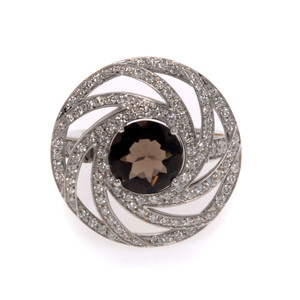 An image of a new Luca Carati women's cocktail ring featuring a large central smoky quartz gemstone surrounded by concentric circles of small round cut diamonds in white gold. The view is a direct overhead shot, showing the ring's intricate design and the sparkle of the gemstones on a neutral background. The distance is close-up, providing a clear view of the ring's details and craftsmanship.
