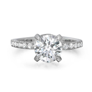 An image of a Rachel Koen brand engagement ring for women, showcasing a prominent round-cut lab grown diamond centered in a four prong setting. The ring, viewed at a close distance and a slight angle, reveals smaller prong set lab grown diamonds along the band, all against a white background.