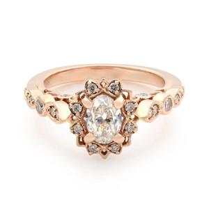 An image of a Rachel Koen brand engagement ring for women, showcasing a central oval-cut diamond set in a rose gold band. The ring is positioned at a slight angle to the camera, allowing for a clear view of the intricate setting and the smaller round diamonds encircling the main stone. The image is taken from a close-up perspective to highlight the ring's details, and the lighting reflects off the diamonds to emphasize their brilliance.