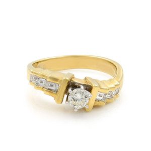 An image of a Rachel Koen women's engagement ring with diamonds, displayed at a three-quarter angle emphasizing the ring's design. The band is made of polished yellow gold, and the ring features a prominent round-cut central diamond held by a gold setting. Flanking the main stone are channel-set princess cut diamonds, adding elegance and sparkle. The image is taken from a medium distance, providing a clear view of the ring's details against a white background.