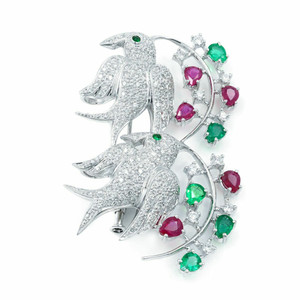 An image of a Rachel Koen women's 18k white gold brooch adorned with diamonds and emerald and ruby gemstones. The brooch features two intricately designed birds positioned diagonally, embellished with sparkling diamonds. The birds are set against a backdrop of a swirling branch adorned with vibrant emerald and ruby gemstones. The image is taken from a close-up angle, highlighting the intricate details and the shimmering quality of the gemstones and diamonds.Excellent pre-owned condition. 