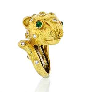 An image of a women's 18k yellow gold ring by David Webb featuring a sculpted lion head design. The ring is adorned with diamonds and emerald, with prominent green emerald eyes. The gold surface is textured, and the diamonds are set throughout the design. Photographed from a side angle at close range, the intricate details and craftsmanship are clearly visible.Excellent pre-owned condition. 