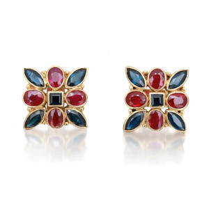 An image of a pair of women's earrings by Rachel Koen, featuring a symmetrical design adorned with ruby and blue sapphire gemstones. The earrings are positioned side by side, facing forward at a slight angle, captured from a close-up distance. The intricate arrangement of oval and marquise-shaped stones is set in a yellow gold frame, highlighting the vibrant colors and elegant craftsmanship.