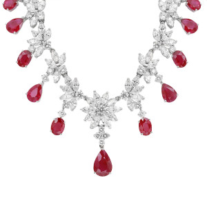 An image of a Rachel Koen women's necklace featuring diamonds and rubies. The necklace is displayed from a front angle at a close distance, highlighting its intricate design in white gold. It showcases a series of diamond clusters, resembling floral patterns, interspersed with large, teardrop-shaped red ruby gemstones. The arrangement creates a symmetrical and elegant appearance. Excellent preowned condition.