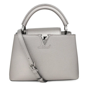 This is an image of a Louis Vuitton Capucines BB Metallic Grey Top Handle Handbag, positioned upright for a front view at a straight angle. This bag showcases a textured Taurillon leather surface in metallic grey, with a prominent "LV" logo in the center. It features a sturdy, curved handle at the top and a diagonally hanging detachable shoulder strap. The handbag includes a single rolled leather reinforced top handle with stylized links and an adjustable, detachable crossbody strap. The crossover flap inserts behind the prominent LV and opens to a partitioned leather interior with a zip pocket. The bag is finished with silver-toned metal hardware and four protective metal bottom studs. This is unworn item appears new but shows some very faint, barely visible scratches and creasing on the leather from storage and handling. It comes with the original box, dust bag, and receipt
