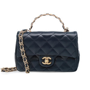 An image image showcases a Chanel 23a Rectangular Top Handle Flap Crossbody handbag in a stunning marine blue, quilted lambskin leather. The bag is presented upright, viewed directly from the front. It features a gold-toned metal chain handle and a striking gold-tone metal logo clasp with a buckle closure. The handbag is equipped with a chain link and leather shoulder strap and a top handle. The interior is lined with leather and includes one zipped pocket while the exterior offers one open pocket. This is a display model, and while it is unworn, there are slight, barely visible scratches on the metal parts and minor creasing on the leather due to handling. The bag comes with the original dust bag, box, and store receipt.