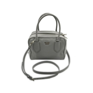 This image presents a Prada Bauletto Ladies Shoulder Handbag in granite gray, crafted from calf leather. The bag is positioned upright and viewed from a slight angle, showcasing its two handles and a long, detachable, and adjustable shoulder strap draped in front. The handbag exhibits a light gray exterior with a unique pistachio interior. It features silver-tone hardware and a top zip closure, leading to an interior pouch with pockets. Crafted in Italy, the bag comes with minor scuffs and scratches from handling. A dust bag is included. 
