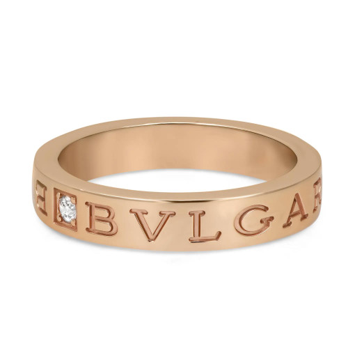 An image of a Bvlgari B.Zero1 unisex adult band ring with diamonds, showcased on a white background. The ring is positioned horizontally in the center with a slight tilt, allowing a clear view of the brand name engraved on the band. The 18k rose gold ring features the letters "BVLGARI" cut out around the circumference with one diamond set between the letters, offering a close-up view that highlights the stone's brilliance and the ring's polished finish. The perspective is a close mid-range shot, focusing on the upper side of the ring and capturing the details at an eye-level angle.