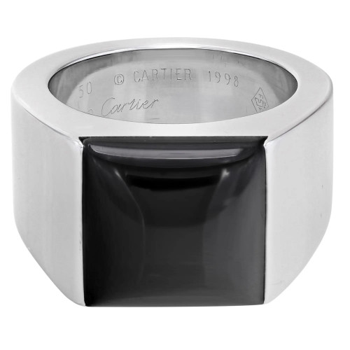 An image of a unisex adult Cartier ring with gemstone, viewed from a top-front angle at close distance. The ring features a prominent black Onyx gemstone set in a thick, polished white gold band with squared edges. The brand's hallmark and serial number are engraved on the interior of the band, partially visible from this perspective. Great preowned condition.
