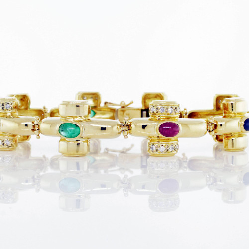 An image of a luxurious Catherine Mullen women's bracelet with diamonds and gemstones. The bracelet is positioned horizontally across the center of the frame, viewed from a close distance, and captured at eye level. It features a series of gold links, each adorned with a central emerald, ruby and sapphire and flanked by smaller diamonds. The polished yellow gold links and sparkling gemstones are crisply focused, casting reflections on the white surface below. Bracelet is in excellent preowned condition.
