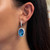 An image of a Rachel Koen earring with diamonds and topaz, modeled on a woman's left ear. The earring features a large oval blue topaz surrounded by a halo of smaller diamonds. It is viewed at a close distance and a slight angle, showcasing the earring's sparkle and intricate design.The condition of the earring is pre owned. 