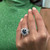 An image of a woman's hand displaying a Rachel Koen ring with diamonds and blue sapphire. The ring features a large central blue sapphire surrounded by a halo of smaller diamonds, set on a 14k white gold band. The hand is positioned against a green textured background, with the ring clearly visible in a close-up frontal view. The fingers are slightly spread, and the ring is worn on the ring finger, with natural lighting accentuating the sparkle of the stones.