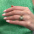 An image of a woman's hand displaying a Rachel Koen brand ring with diamonds and green emeralds, viewed from a close, slightly above angle, emphasizing the details and design of the jewelry piece. The ring is worn on the ring finger, showcasing a central diamond surrounded by green emeralds set in a 14k yellow gold band against a textured green background.