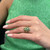 An image of a woman's hand with pale pink manicured nails, showcasing a Rachel Koen ring with green emeralds. The ring is worn on the ring finger and features multiple small green emeralds set in a 14k yellow gold band with a honeycomb-like design. The hand is positioned against a green fabric background with a subtle circular texture. The photo is taken from a close, slightly above angle, focusing on the ring to capture the intricate details and the shine of the gemstones.