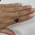 An image of a Rachel Koen pendant necklace with diamonds and Ruby presented on a hand, showcasing a close-up view. The necklace features a large, oval-cut red Ruby encircled by smaller diamonds set in a halo design. The pendant is centered and suspended from a delicate white gold chain, which drapes over the fingers at a slight diagonal angle. The hand is resting against a white textured background, providing contrast that emphasizes the sparkling jewelry. The photo is taken from a top-down perspective at a close distance, allowing for clear visibility of the necklace's details.