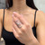 An image of a Messika brand diamond ring worn on the ring finger of a woman's left hand, prominently displayed at the center with a slight tilt towards the viewer. The ring features multiple diamond-encrusted bands interlocking in a complex, elegant design. The woman is shown from a close distance, with her hand positioned against a blurred background to emphasize the ring.