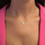 An image of a Messika brand necklace with diamonds, designed for women, displayed on a model. The necklace features a delicate chain with multiple diamonds spaced along its length, culminating in a larger, triangular diamond pendant at the center. The pendant sits just above the collarbone, with the model wearing a pink garment. The image is a close-up, taken from a front-facing angle.