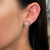 An image of a Messika brand earring with diamonds, designed for women, as worn on the left ear. The earring features a prominent circular design with a central diamond cluster and is viewed from a side angle, showcasing its sparkle and position against the earlobe. The photograph is a close-up, providing a clear view of the earring's intricate details and the reflective quality of the diamonds.