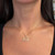 An image of a Messika brand necklace with diamonds, worn by a person positioned centrally in the frame. The necklace features a delicate gold chain with small diamond embellishments and a larger diamond-studded pendant in the shape of a stylized design. The photo is taken at a close distance, showing the necklace from a straight-on angle against the wearer's neck and upper chest area, highlighting the sparkle and design of the jewelry. 