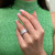 An image of a woman's hand with a manicured pink nail polish, positioned at a three-quarter angle, displaying a Messika diamond ring. The ring is worn on the ring finger, prominently featuring multiple rows of small, sparkling diamonds set in a wide band. The image is taken from a close distance, focusing on the ring against the backdrop of a green textured garment worn by the person.