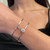 An image of a Messika women's bracelet with diamonds, displayed on a person's wrist. The bracelet features a central circular diamond setting and a slender, open-ended band. The photo is taken at a close distance, showcasing the bracelet in clear detail against the skin. The bracelet is positioned roughly at the center of the frame with a slight tilt, allowing a good view of its sparkling diamonds and elegant design. The person's attire appears to be black, providing a contrasting background that accentuates the jewelry's shine.