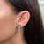 An image of a Messika brand earring with diamonds, modeled on a woman's left ear. The earring is a vertical bar design, featuring a row of sparkling diamonds set in a polished white gold frame. The photograph is a close-up side view, with the earring positioned centrally, providing a clear view of the earring's design and the diamonds' brilliance. The background shows the woman's straight dark hair, partially obscuring her neck and shoulder.