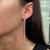 An image of a woman's ear wearing a Messika earring with diamonds. The earring is a long, slender, vertical drop style adorned with sparkling diamonds, and features a few larger diamonds interspersed along its length, creating a luxurious gradient effect. The photo is a close-up, side view, focusing on the earring as it dangles elegantly from the wearer's lobe against her dark hair and fair skin. The background is softly blurred, emphasizing the jewelry's detail and brilliance.