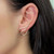An image of a Messika earring for women, featuring a curved design with multiple diamonds embedded throughout, worn on the left ear. The earring has a serpentine shape that contours along the edge of the ear, with the camera angle capturing the piece in a close-up side view, highlighting the sparkle and intricate setting of the diamonds.