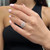 An image of a Messika women's ring with diamonds, displayed on a hand with light pink manicured nails. The ring is shown in close-up from a slightly elevated angle, focusing on the intricate design and sparkling stones. The hand is positioned at a relaxed angle with the fingers slightly spread, giving a clear view of the ring and its details, which include multiple bands and a prominent diamond setting. The background is blurred, emphasizing the ring as the central subject of the photo.