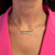 An image of a Messika brand necklace with diamonds, worn by a woman. The necklace features a horizontal bar pendant on a delicate rose gold chain, centered at the base of the neck. The pendant is adorned with a row of sparkling diamonds set against a gold backdrop. The image is a close-up, taken from a front angle, focusing on the necklace against the backdrop of a pink garment. The distance is such that the necklace and upper chest area are clearly visible, with a slight shadow beneath the pendant accentuating its position on the neck.