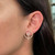 An image of a Messika brand earring with diamonds, designed for women. The earring is showcased on an ear from a side view, close-up angle, highlighting the intricate design which features a circular shape with a smaller circle and a line of diamonds within the larger glittering diamond-encrusted ring. The earring is positioned against the background of the wearer's smooth skin and dark hair, providing a clear and detailed view of the jewelry's sparkle and design.